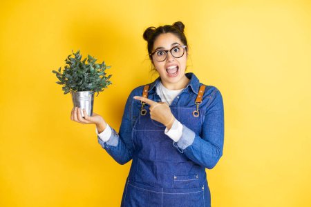 Photo for Young caucasian gardener woman holding a plant isolated on yellow background surprised, showing and pointing the plant that is on her hand - Royalty Free Image