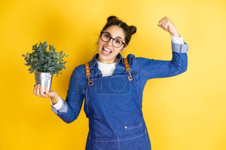 Photo for Young caucasian gardener woman holding a plant isolated on yellow background showing arms muscles smiling proud - Royalty Free Image