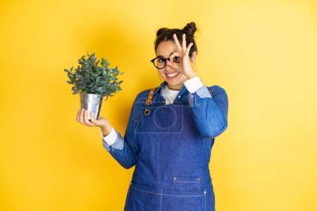 Photo for Young caucasian gardener woman holding a plant isolated on yellow background doing ok gesture shocked with smiling face, eye looking through fingers - Royalty Free Image