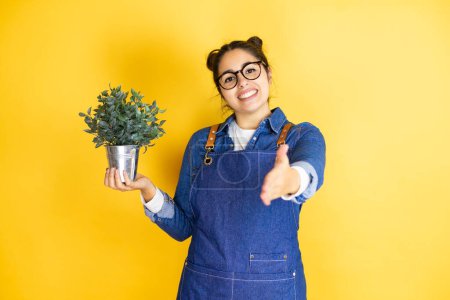 Photo for Young caucasian gardener woman holding a plant isolated on yellow background smiling friendly offering handshake as greeting and welcoming - Royalty Free Image