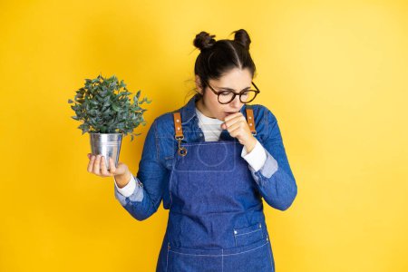 Photo for Young caucasian gardener woman holding a plant isolated on yellow background with her hand to her mouth because she's coughing - Royalty Free Image