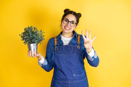 Photo for Young caucasian gardener woman holding a plant isolated on yellow background showing and pointing up with fingers number four while smiling confident and happy - Royalty Free Image