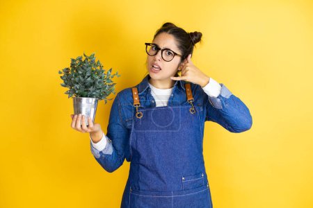 Photo for Young caucasian gardener woman holding a plant isolated on yellow background confused doing phone gesture with hand and fingers like talking on the telephone - Royalty Free Image