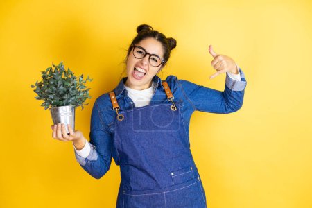 Photo for Young caucasian gardener woman holding a plant isolated on yellow background shouting with crazy expression doing rock symbol with hands up - Royalty Free Image
