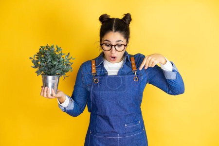 Photo for Young caucasian gardener woman holding a plant isolated on yellow background surprised, looking down and pointing down with fingers and raised arms - Royalty Free Image