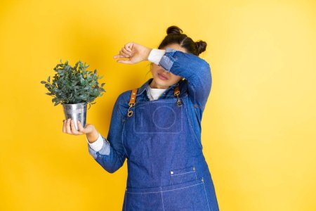 Photo for Young caucasian gardener woman holding a plant isolated on yellow background covering eyes with arm smiling cheerful and funny. Blind concept. - Royalty Free Image