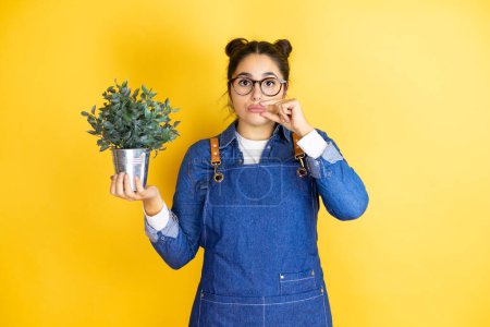 Photo for Young caucasian gardener woman holding a plant isolated on yellow background mouth and lips shut as zip with fingers. Secret and silent, taboo talking - Royalty Free Image