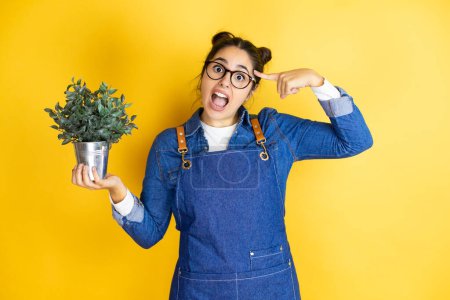 Photo for Young caucasian gardener woman holding a plant isolated on yellow background smiling and thinking with her fingers on her head that she has an idea. - Royalty Free Image