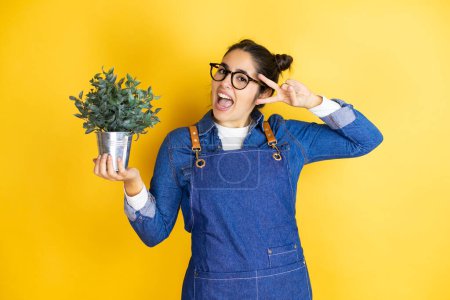 Photo for Young caucasian gardener woman holding a plant isolated on yellow background Doing peace symbol with fingers over face, smiling cheerful showing victory - Royalty Free Image