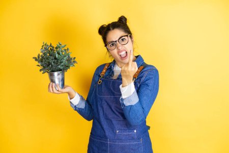 Photo for Young caucasian gardener woman holding a plant isolated on yellow background showing middle finger doing fuck you bad expression, provocation and rude attitude. screaming excited - Royalty Free Image