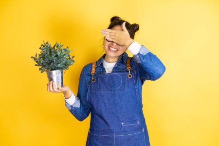 Photo for Young caucasian gardener woman holding a plant isolated on yellow background covering eyes with hands smiling cheerful and funny. Blind concept. - Royalty Free Image