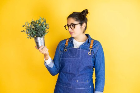 Photo for Young caucasian gardener woman holding a plant isolated on yellow background relaxed with serious expression on face. simple and natural looking the plant - Royalty Free Image