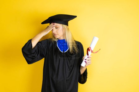 Beautiful blonde young woman wearing graduation cap and ceremony robe putting one hand on her head smiling like she had forgotten something