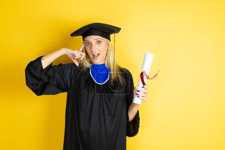 Beautiful blonde young woman wearing graduation cap and ceremony robe smiling and thinking with her fingers on her head that she has an idea.