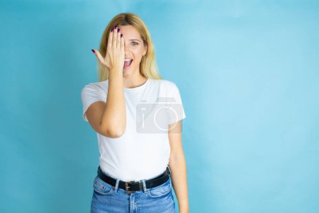 Young beautiful woman wearing casual t-shirt over isolated blue background covering one eye with hand, confident smile on face and surprise emotion.