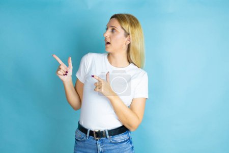 Junge schöne Frau in lässigem T-Shirt vor isoliertem blauen Hintergrund überrascht und zeigt mit den Fingern zur Seite