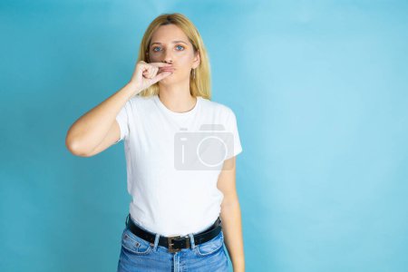 Young beautiful woman wearing casual t-shirt over isolated blue background mouth and lips shut as zip with fingers. Secret and silent, taboo talking