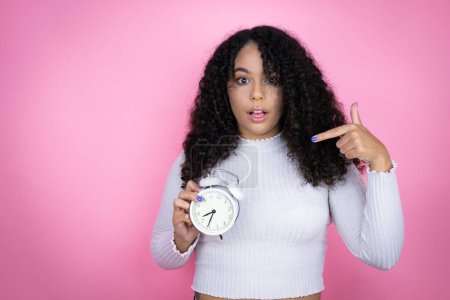 Afroamerikanerin in lässigem Pullover vor rosa Hintergrund überrascht hält und zeigt eine Uhr