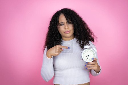 African american woman wearing casual sweater over pink background serious holding and pointing a clock