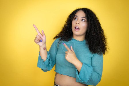 Afroamerikanerin in lässigem Pullover vor gelbem Hintergrund überrascht und zeigt mit den Fingern auf die Seite