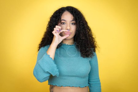 African american woman wearing casual sweater over yellow background mouth and lips shut as zip with fingers. Secret and silent, taboo talking