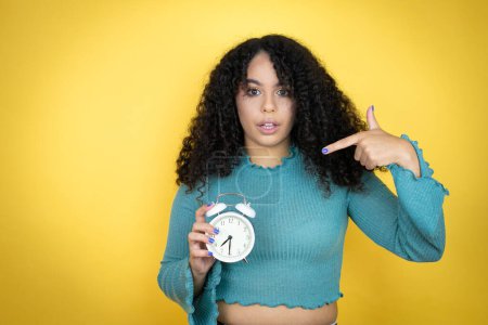 Afroamerikanerin in lässigem Pullover vor gelbem Hintergrund überrascht, als sie eine Uhr hält und zeigt
