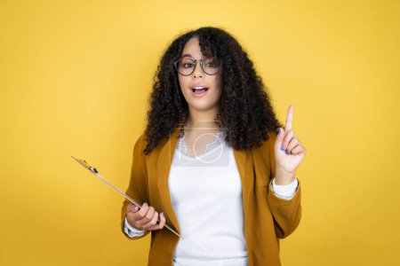 Photo for African american business woman with paperwork in hands over yellow background surprised and thinking with her finger on her head that she has an idea. - Royalty Free Image