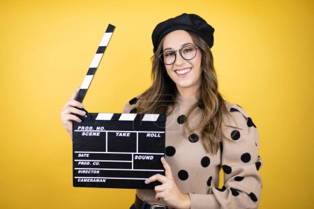 Young beautiful brunette woman wearing french beret and glasses over yellow background holding clapperboard very happy having fun