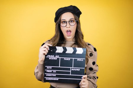 Young beautiful brunette woman wearing french beret and glasses over yellow background holding clapperboard very happy having fun