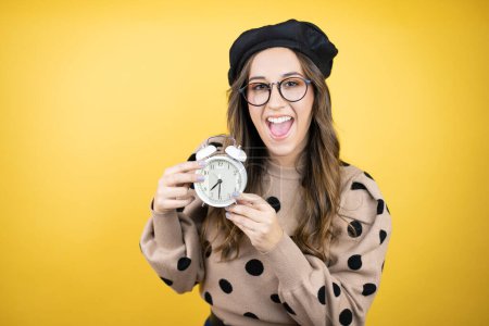 Young beautiful brunette woman wearing french beret and glasses over yellow background serious holding and pointing a clock