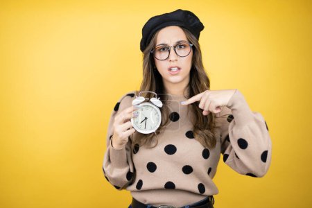 Jeune belle femme brune portant un béret français et des lunettes sur fond jaune tenue sérieuse et pointant une horloge