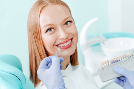 Téléchargez les photos : Jolie jeune femme avec des dents blanches naturelles dans la clinique dentaire. Mains médecin dentiste avec palette de couleurs des dents à côté par le visage. Sourire dents saines concept - en image libre de droit