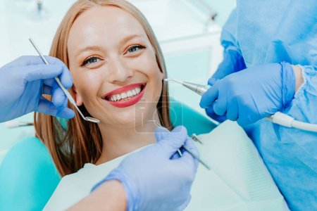 Téléchargez les photos : Attrayant jeune femme smilling avec des dents blanches naturelles dans la clinique dentaire. Mains médecin dentiste avec des outils médicaux. Concept de dents saines - en image libre de droit