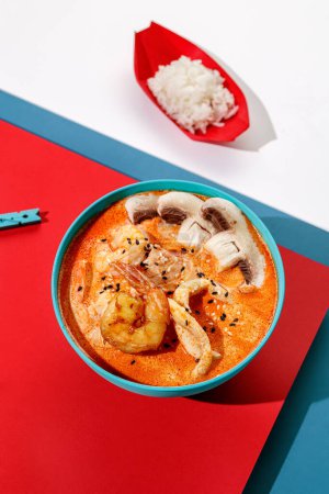 Foto de Sopa de Tom yum con camarones y arroz en boul de color pastel azul en fondo azul y rojo con luz solar y sombras, minimalismo, vista superior. - Imagen libre de derechos
