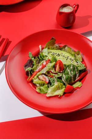 Photo for Fresh vegan green salad with avocado, tomatoes , red bell peper and nut sauce in red color boul at red and white background with sunlight and shadows, minimalism. - Royalty Free Image