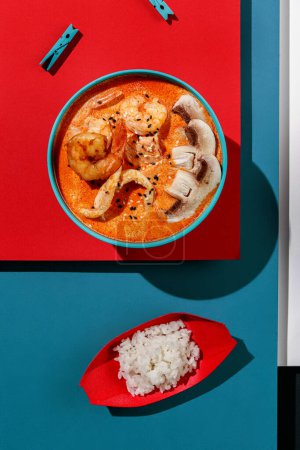 Foto de Tom yum soup with shrimps and rice in blue pastel color boul at blue and red background with sunlight and shadows, minimalism,top view. - Imagen libre de derechos