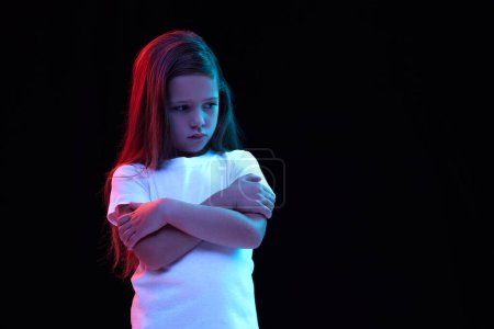 Photo for Resentment. Sad little girl, kid with long dark hair wearing white t-shirt expressing emotions isolated over black background. Concept of children emotions, fashion, beauty, back to school and ad - Royalty Free Image
