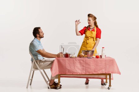 Foto de Retrato de una hermosa mujer cocinando, horneando y hombre sentado en la mesa aislado sobre fondo blanco. Comestibles. Concepto de estilo retro, deberes domésticos, moda antigua, estilo de vida. Copiar espacio para anuncio - Imagen libre de derechos
