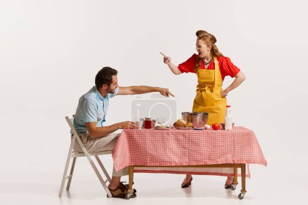Foto de Retrato de pareja joven y emotiva peleando mientras desayunaba aislado sobre fondo blanco. Mujer cocinando, hombre comiendo. Concepto de estilo retro, deberes domésticos, moda antigua, estilo de vida. Copiar espacio para anuncio - Imagen libre de derechos