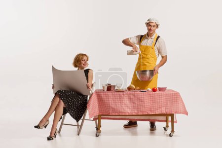 Foto de Hombre alegre en delantal cocinando mientras mujer sentada en la mesa y leyendo periódico aislado sobre fondo blanco. Concepto de estilo retro, deberes domésticos, moda antigua, estilo de vida. Copiar espacio para anuncio - Imagen libre de derechos