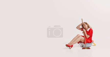 Foto de Mujer elegante sentada en un tazón con ropa y sujetando la fregona aislada sobre fondo blanco. Cansado y agotado. Concepto de estilo retro, deberes domésticos, moda antigua, estilo de vida. Copiar espacio para anuncio - Imagen libre de derechos