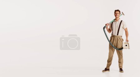 Foto de Retrato de un joven guapo y elegante posando con una aspiradora vintage aislada sobre fondo blanco. Concepto de estilo retro, deberes domésticos, moda antigua, estilo de vida. Copiar espacio para anuncio - Imagen libre de derechos