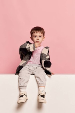 Portrait of cute little toddler boy sitting on big box and looking at camera. Emotions, kids fashion, happy childhood concept. Looks calm and sad. Copy space for ad