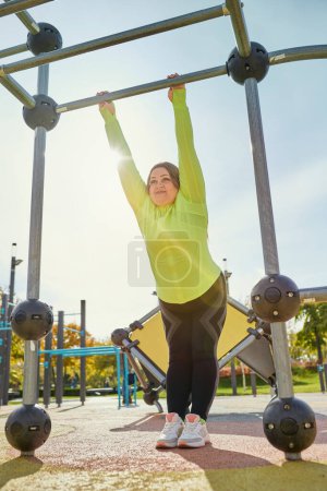 Photo for Pull-ups on the bar. Young woman in colorful sportswear doing fitness exercises at street public sports ground, outdoors. Sunny autumn day. Concept of wellness, sport, health, mood, body positive - Royalty Free Image