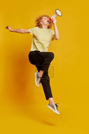 Photo for News. Full-length portrait of active young red-haired girl jumping with megaphone isolated on yellow studio background. Human emotions, facial expression, beauty concept. Copy space for ad - Royalty Free Image