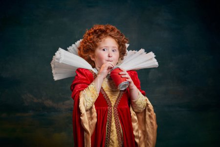 Photo for Portrait of little red-headed girl in costume of royal person drinking soda isolated over dark green background. Concept of historical remake, comparison of eras, medieval fashion, emotions, queen - Royalty Free Image