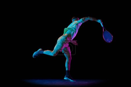 Téléchargez les photos : Énergie et pouvoir. Portrait dynamique d'un joueur de tennis masculin énergique en tenue de sport jouant au tennis isolé sur fond sombre sous la lumière du néon. Concept de mouvement, vitesse, sport professionnel. - en image libre de droit