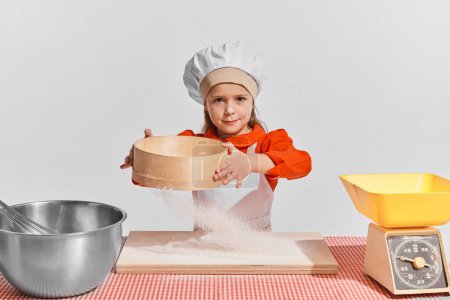 Photo for Cute little child, girl in image of chef cooking over grey background. Sifting flour. Concept of childhood, creativity, retro style, vintage fashion, future profession, friendship, art - Royalty Free Image