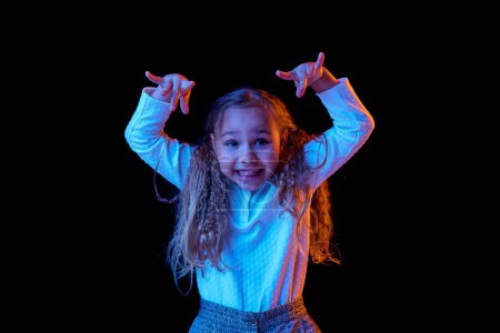 Photo for Resentment. Funny little girl, kid with long hair wearing white t-shirt expressing emotions isolated over black background in neon light. Concept of children emotions, fashion, beauty, back to school - Royalty Free Image