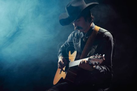 Photo for Portrait of man with moustaches in country style clothes playing guitar isolated over dark blue background with smoke. Making music. Concept of music, creativity, inspiration, hobby, lifestyle - Royalty Free Image